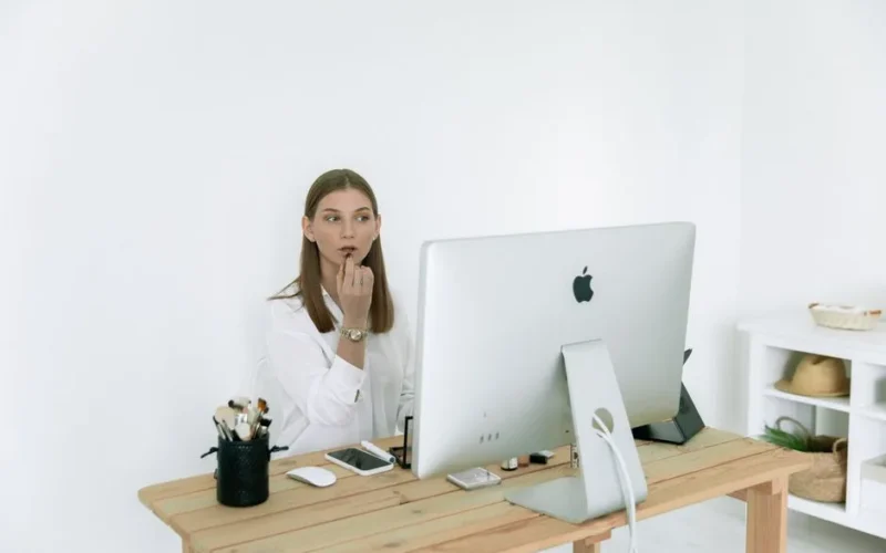 Woman applying makeup at desk, balancing work and beauty routine indoors.