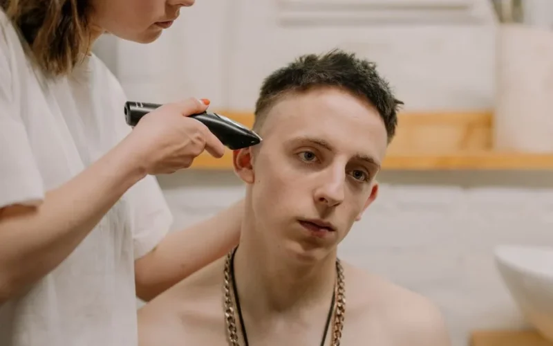 Caucasian young man receiving a haircut at home, trimmed by an adult woman using an electric trimmer.