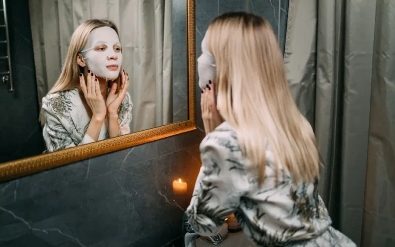 A woman applies a sheet mask in front of a mirror, enjoying a serene skincare routine.
