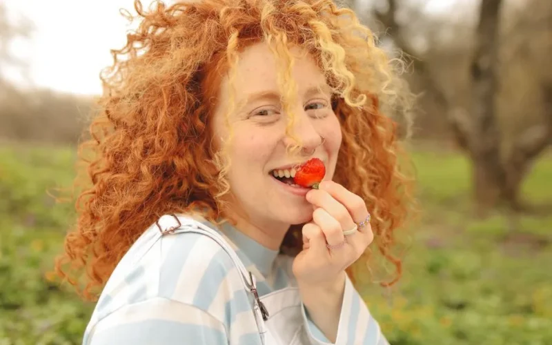 Smiling woman with curly red hair eating a strawberry in a sunny outdoor setting.