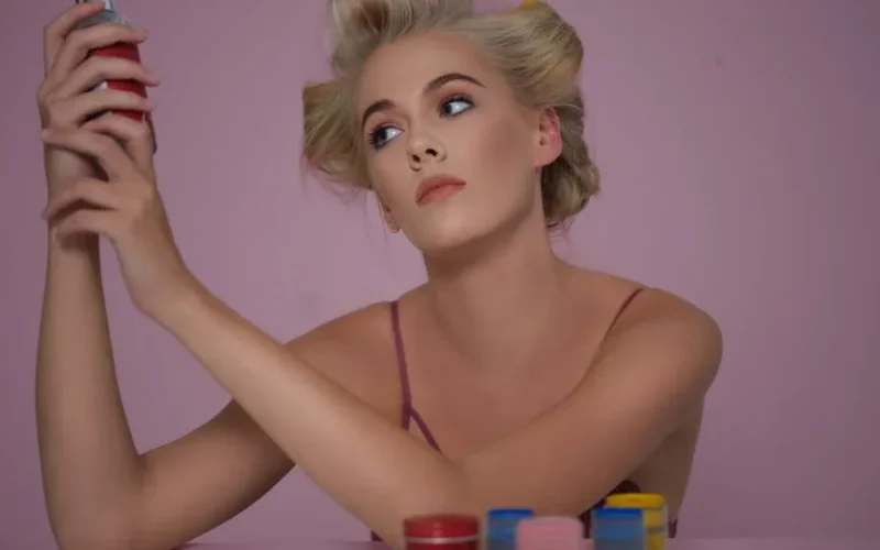 Portrait of a woman with styled hair holding a can in a studio setting, surrounded by colorful hair products.