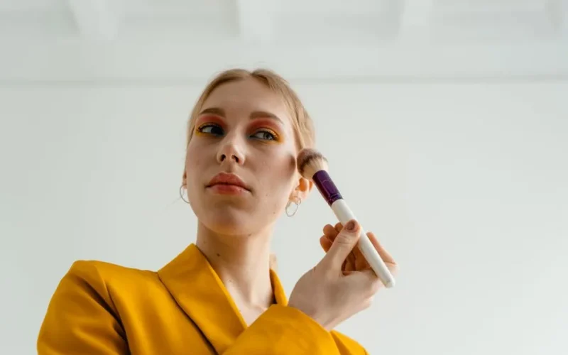 Woman in yellow attire applying blush with a makeup brush indoors.