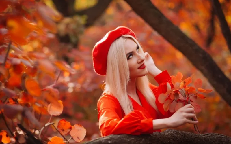 Blonde woman in red beret surrounded by vibrant autumn leaves in a forest setting.