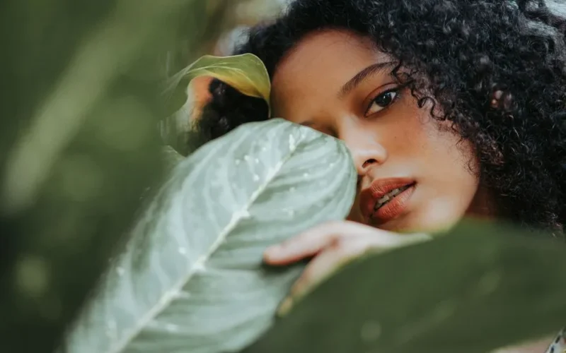 Beautiful serene ethnic female with black curly hair standing in garden and touching green plant leaves while looking at camera