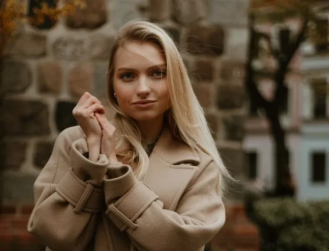 A stylish woman in a beige coat poses confidently outdoors with a stone wall backdrop.