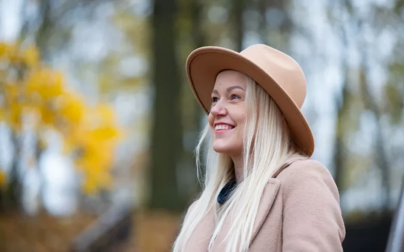 Blonde woman smiling outdoors in fall wearing a beige hat and coat.