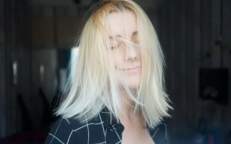 Smiling young female with messy blond hair in casual clothes standing in room with closed eyes