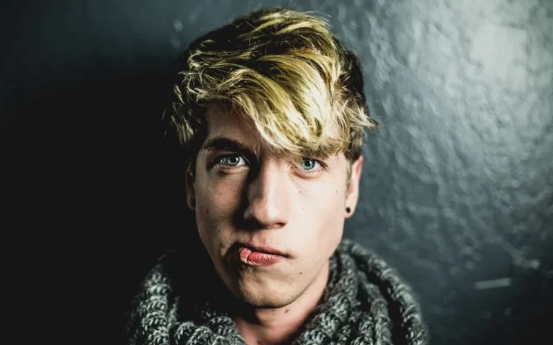Portrait of a young man with blonde hair wearing a scarf against a dark background.