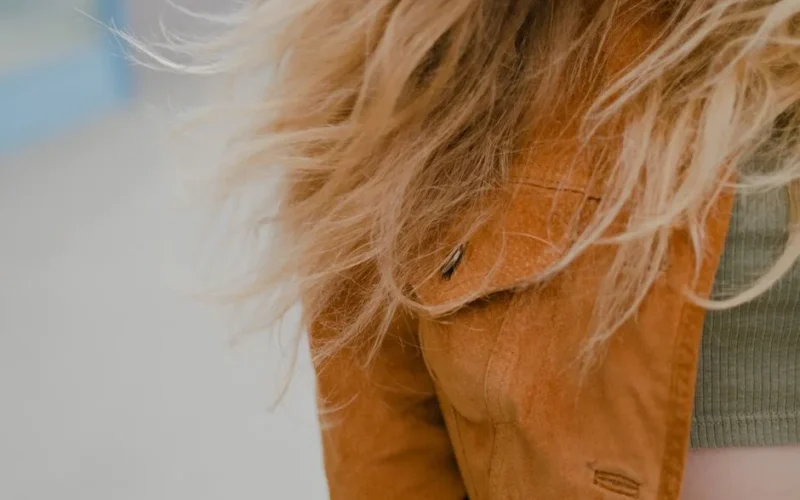 Close-up image focusing on flowing blonde hair and a brown jacket, outdoors.