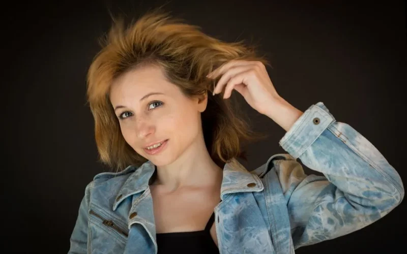 Close-up portrait of a smiling woman with blonde hair in a denim jacket holding her hair, studio shot.