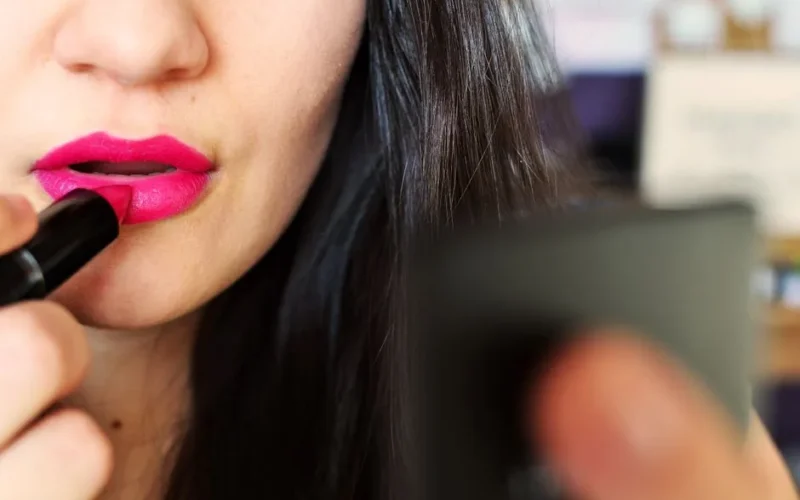 Close-up of a woman applying bright pink lipstick while looking in a mirror, highlighting beauty and makeup.