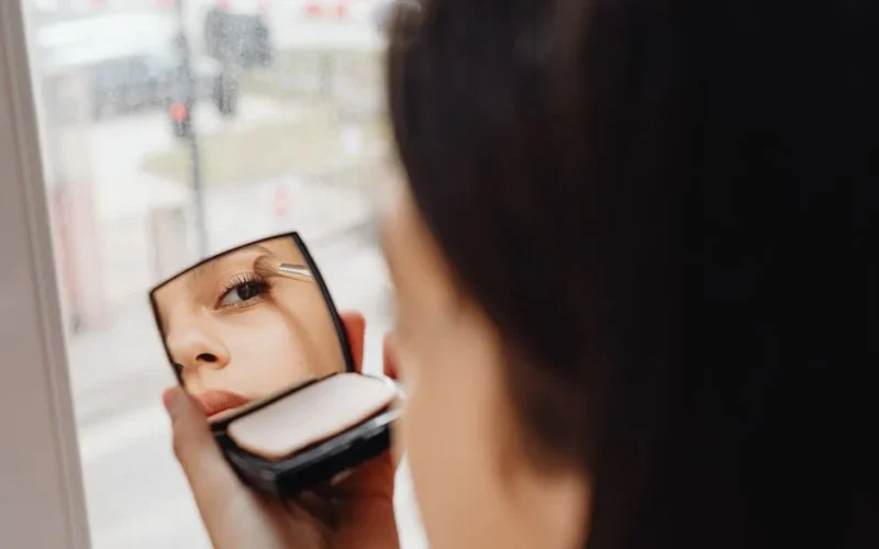 A woman applies eye makeup using a compact mirror, showcasing a close-up reflection effect indoors.