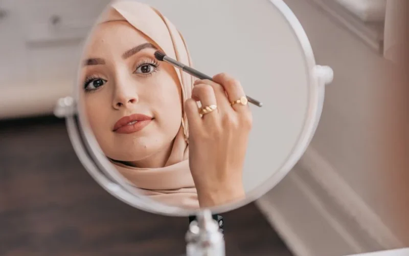 Close-up of a woman applying makeup with a brush, reflecting in a round mirror indoors.