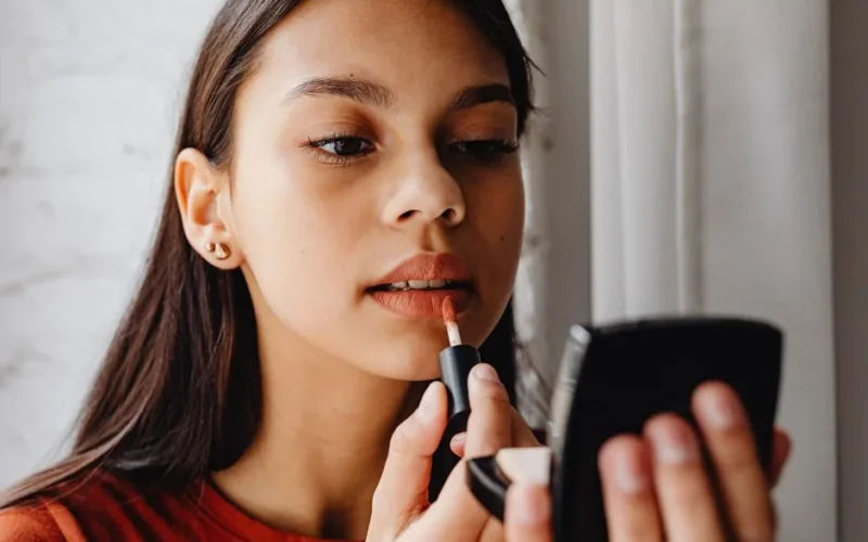A young woman applies lipstick while looking in a handheld mirror.