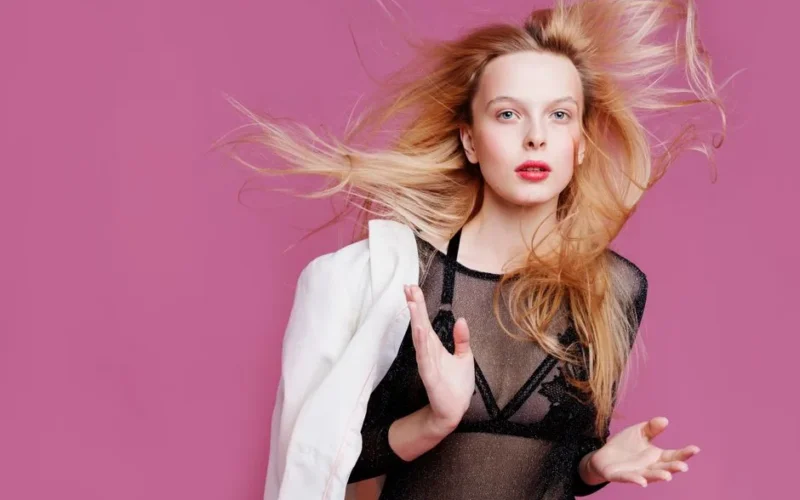 Elegant portrait of a blonde woman in a stylish outfit against a pink background.
