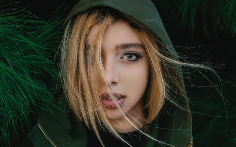 Close-up portrait of a woman with blonde hair and green hood, surrounded by foliage.