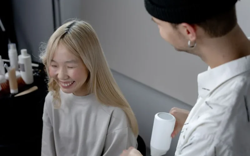 A hairstylist using a blow dryer on a smiling Asian woman in a hair salon.