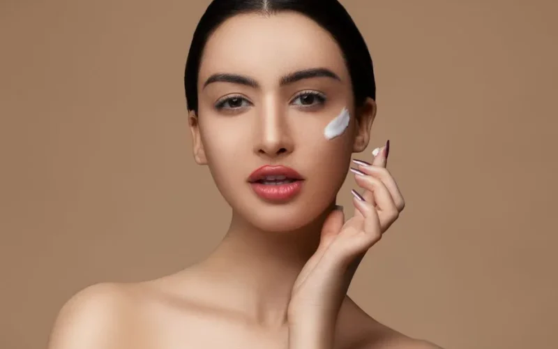 Crop sensitive female with makeup and red lips applying cream on delicate face skin while looking at camera on beige background