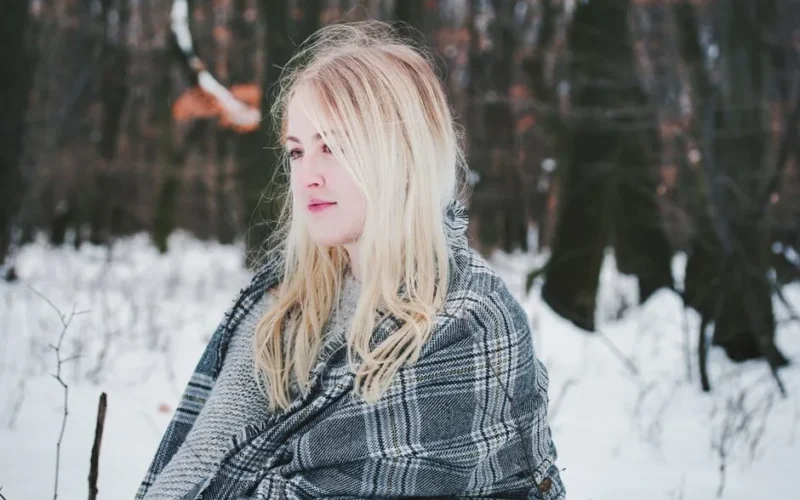 A blonde woman wrapped in a plaid scarf in a snowy winter forest setting. Serene and contemplative mood.