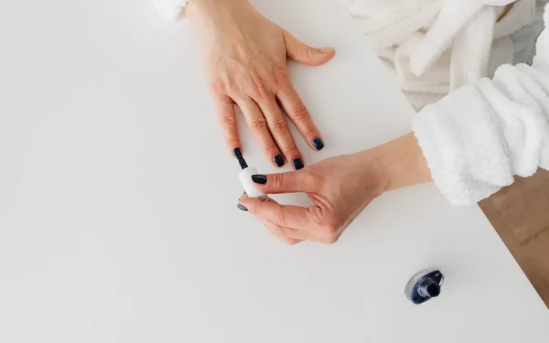 Top view of a woman painting her nails with dark polish, wearing a cozy robe.