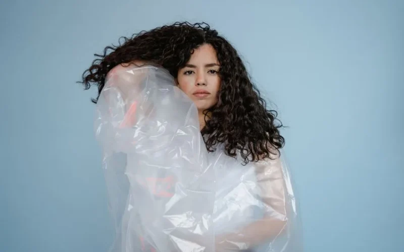A woman with curly hair wrapped in transparent plastic against a blue background.