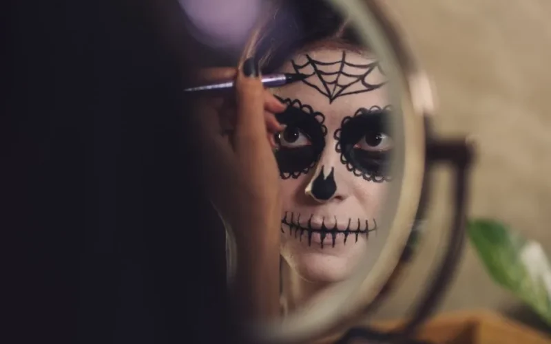Woman in front of mirror applying Day of the Dead face paint for Halloween celebration.