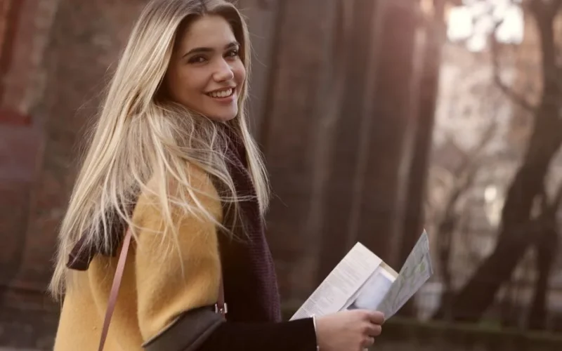 Young blonde woman smiling outdoors, holding a map on a sunny day, enjoying a casual outing.