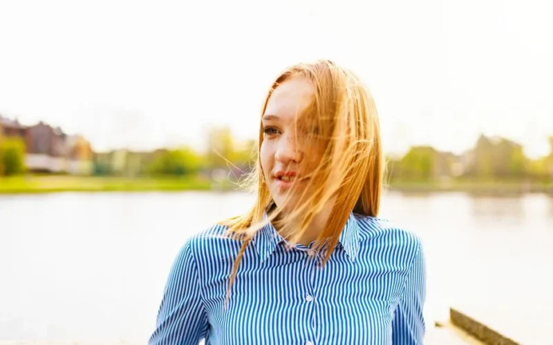 Portrait of a young woman with blonde hair smiling by a lake on a sunny day.