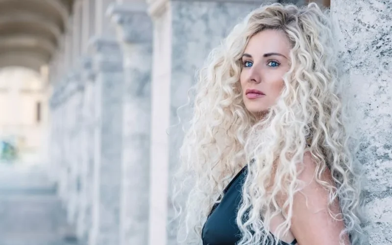 Stylish outdoor portrait of a woman with curly blonde hair leaning against a stone column.