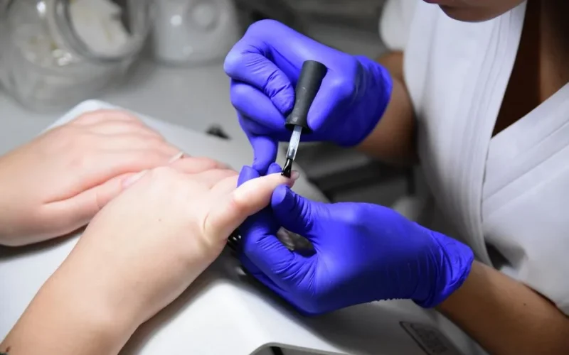 Close-up of a manicurist applying nail polish wearing purple gloves in a salon.