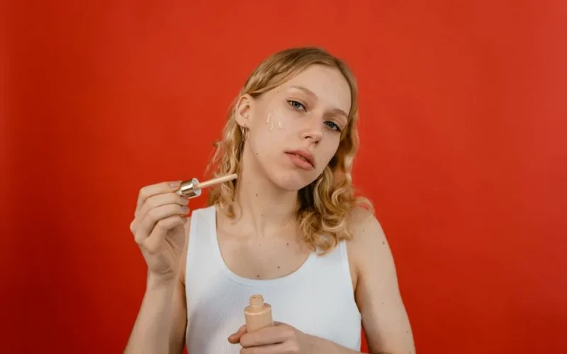 A young woman in a tank top applies concealer to her face with a dropper against a red background.