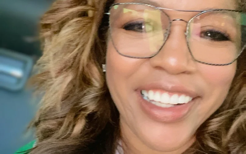 Portrait of Danyel Smith smiling and wearing glasses. Smith is a woman with long sandy brown hair with highlights and is wearing a white shirt with green outer layer.