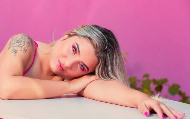 A woman with blonde hair, tattoos, and vibrant makeup resting on a table against a pink background.