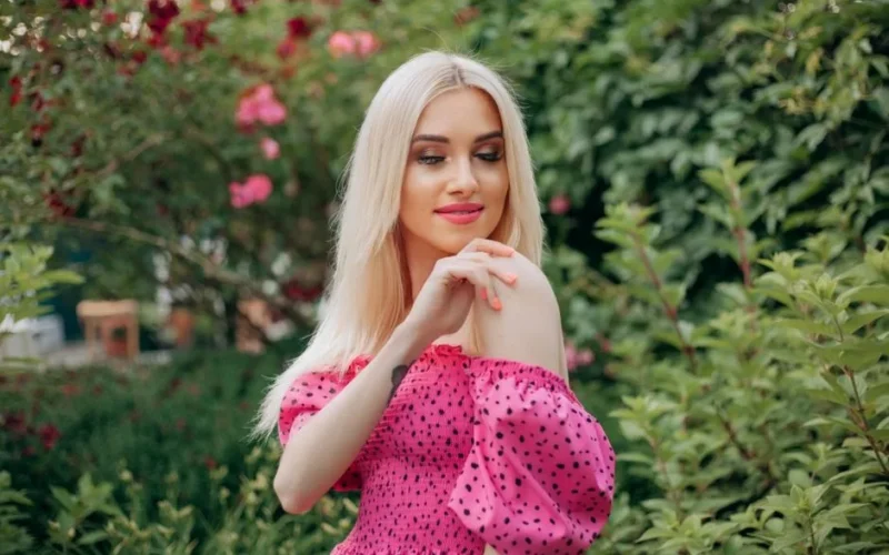 Charming portrait of a blonde woman in a pink polka dot dress standing in a lush green garden, smiling softly.