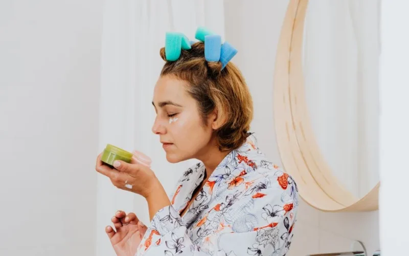 Woman with hair curlers applies skincare cream in front of a bathroom mirror.