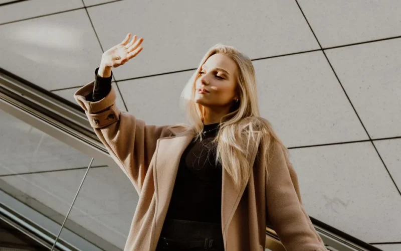 A blonde woman elegantly poses in a wool winter coat, enjoying a sunny day outdoors.