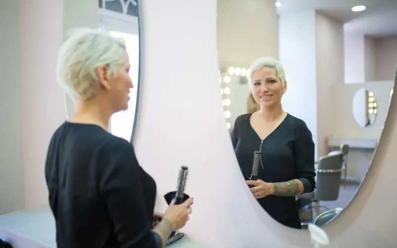 A woman hairdresser smiles in the mirror of a modern salon, holding a brush and hair tools.
