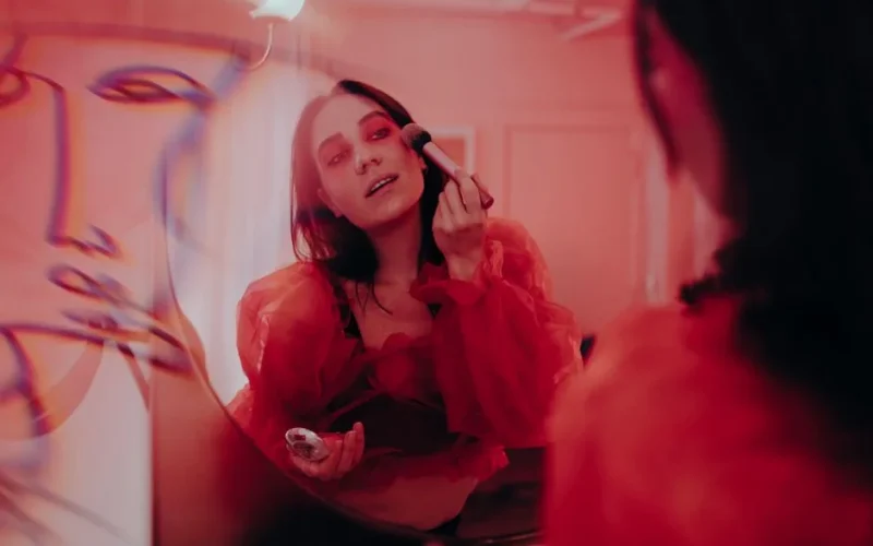 A woman applies makeup in front of a mirror reflecting an artistic red ambiance, creating a stylish, creative scene.