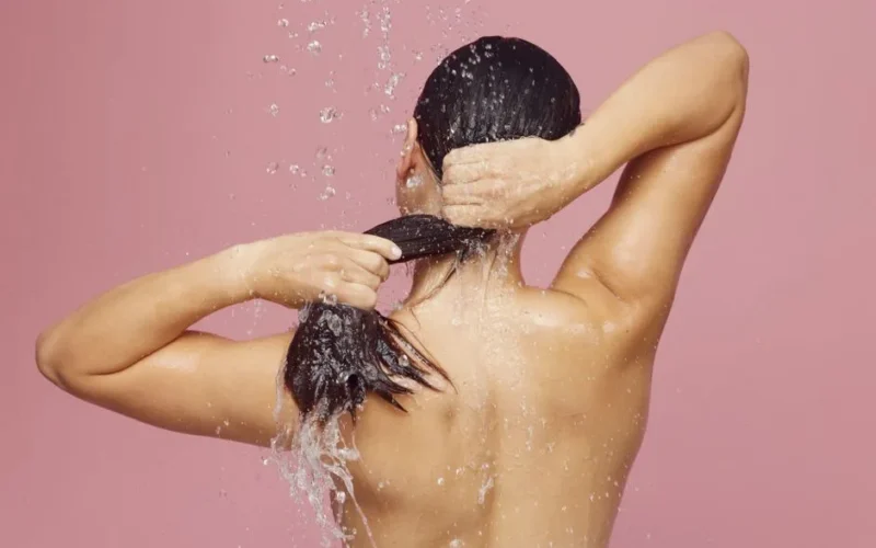 Woman washing long hair under shower with water droplets against a pink background.