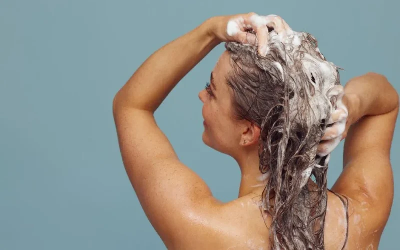 Woman with long hair washing it with shampoo, creating lather, in indoor setting against blue background.