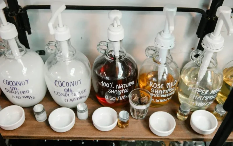 Collection of big dispensers of various multicolored shampoo placed near glass jars and white bowls on wooden shelf