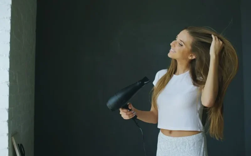 Young woman smiling while using a hairdryer for hair styling indoors.