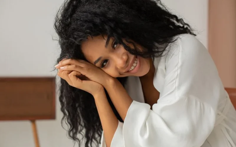 A cheerful woman with curly hair smiling warmly in a white bathrobe, indoors.
