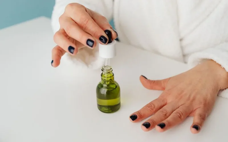 Manicured hands using a dropper to dispense green liquid from a glass bottle.